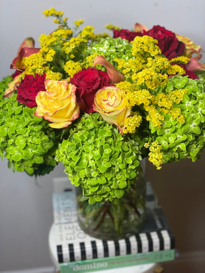 A floral arrangement featuring a variety of flowers including yellow and red roses, and green foliage, presented in a clear glass vase.